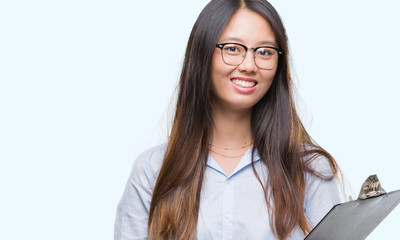 Young asian business woman holding clipboard over isolated background with a happy face standing...