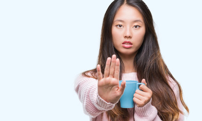 Young asian woman drinking coffee over isolated background with open hand doing stop sign with serious and confident expression, defense gesture