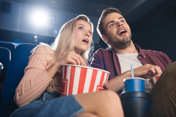 shocked couple with popcorn and soda drink watching film together in cinema