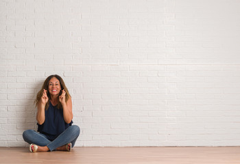 Middle age hispanic woman sitting on the floor over white brick wall smiling crossing fingers with hope and eyes closed. Luck and superstitious concept.