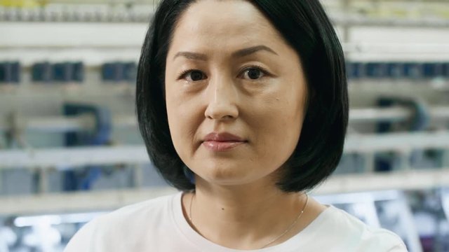Close-up of mid-aged asian woman looking at camera while textile machine processing fabric in background