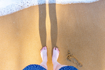 A young pale womens feet standing on the sand with a wave coming wearing spotty shorts