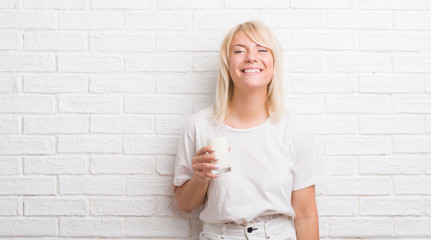 Adult caucasian woman over white brick wall drinking glass of milk with a happy face standing and smiling with a confident smile showing teeth