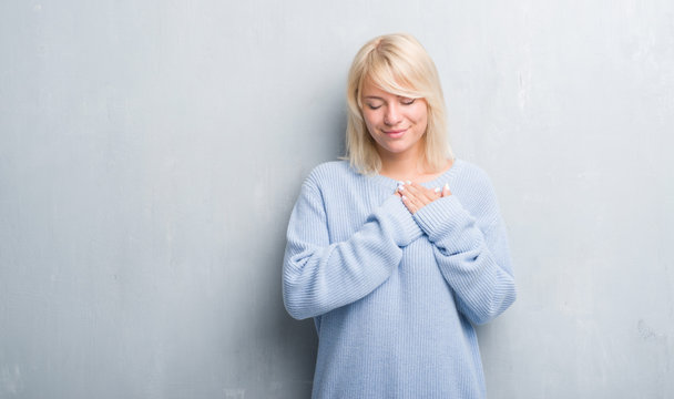 Adult caucasian woman over grunge grey wall wearing winter sweater smiling with hands on chest with closed eyes and grateful gesture on face. Health concept.
