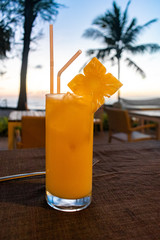 An orange cocktail in a long glass with a pineapple slice on the side, and a sunset and palm trees in the background, tropical