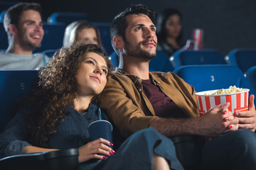 young couple with popcorn holding hands while watching movie together in cinema - obrazy, fototapety, plakaty