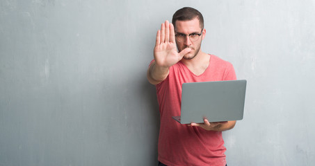 Young caucasian man over grey grunge wall using computer laptop with open hand doing stop sign with serious and confident expression, defense gesture