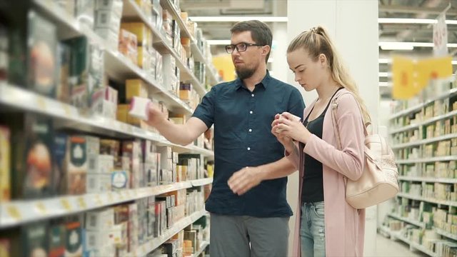Man and woman standing in the aisle with tea, difficult choice. Plenty tea boxes on shelf in supermarket, family couple shopping together.
