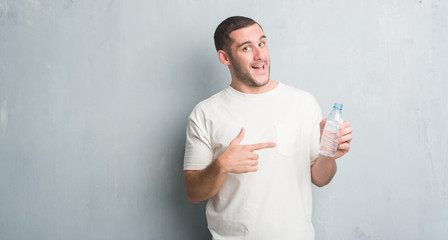 Young caucasian man over grey grunge wall holding bottle of water very happy pointing with hand and finger