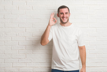 Young caucasian man standing over white brick wall smiling positive doing ok sign with hand and fingers. Successful expression.