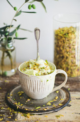 Homemade pistachio ice cream in ceramic mug with green pistachio nuts on rustic wooden kitchen counter, vertical composition. Summer refreshing ice cream dessert