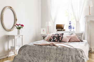 English style bedroom interior featuring a bed with light gray bedding and lemonade pink and rose pattern pillows and white ornamented furniture and decorations. Real photo