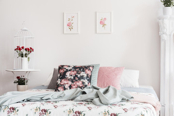 Close-up of flower pattern bed cover and pillows on a bed which is standing against a light color wall with rose illustrations in a feminine classic bedroom interior. Real photo.
