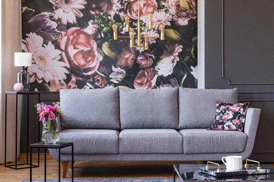 Flowers on black table and grey sofa in living room interior with lamp and wallpaper. Real photo