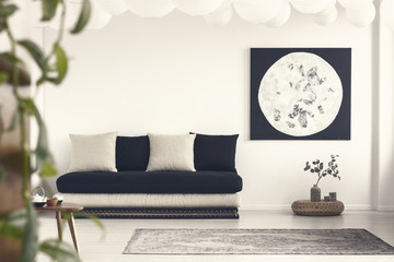 Moon poster above plant on pouf in white living room interior with pillows on black sofa. Real photo