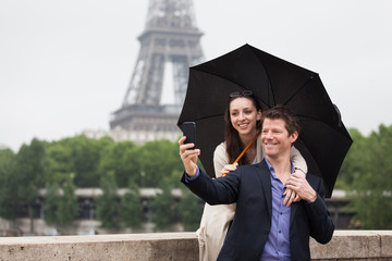 A Smiling Young Couple Take a Selfie Under an Umbrella with the Eiffel Tower behind Them