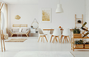 White chairs at dining table near plants in apartment interior with gold poster and beige sofa. Real photo