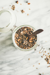 Healthy breakfast preparation. Buckwheat and chocolate granola with hazelnuts in jar with spoon over light marble background, top view, copy space. Clean eating, vegan, vegetarian food concept