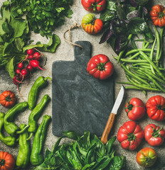 Healthy seasonal food cooking background. Flat-lay of vegetables and greens over concrete background, black marble chopping board in center, top view, copy space. Clean eating, vegan food preparation