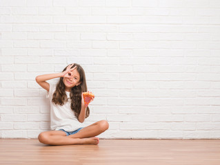 Young hispanic kid sitting on the floor over white brick wall eating pizza slice with happy face smiling doing ok sign with hand on eye looking through fingers