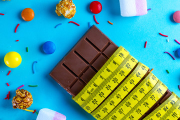 Diet concept, loss weight concept. Chocolate is wrapped in a measuring tape on a blue background with different sweets, candies. Calorie, carbohydrate food.