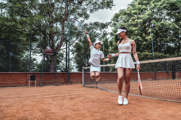 Obraz premium Little champion on tennis court! Full length image of jumping child tennis player with sporty trainer. Tennis coach holds hand of little girl celebrating victory. Active leisure outdoors.