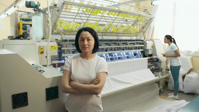 Mid-aged asian woman standing in textile factory and looking at camera while her young colleague using digital tablet and operating textile machine