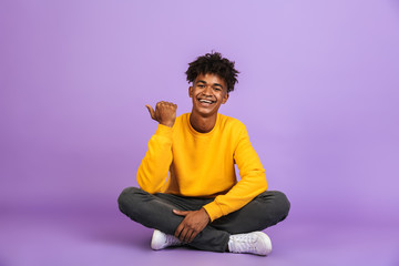 Portrait of cheerful american boy smiling and pointing finger aside at copyspace, while sitting on...