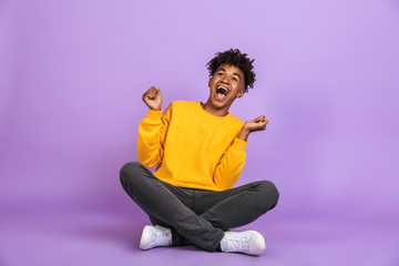 Portrait of excited african american boy screaming and looking aside while sitting on floor with legs crossed, isolated over violet background