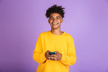 Portrait of teenager boy with afro hairdo smiling and holding smartphone, wearing bluetooth...