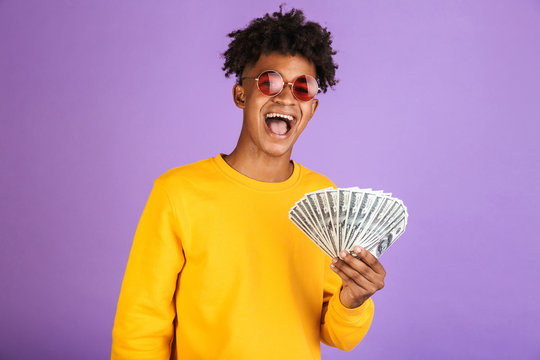 Portrait of fashion african american man wearing sunglasses smiling while holding cash money dollars, isolated over violet background