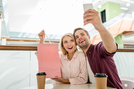 young man taking selfie with girlfriend showing shopping bag at table with disposable cups of coffee in cafe