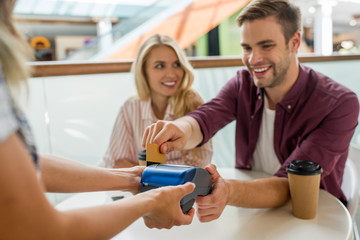 selective focus young man paying by credit card on terminal while his girlfriend sitting near at table with coffee cups in cafe
