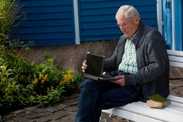 Fototapeta premium schlanker Senior in Freizeitkleidung mit Strickjacke, Jeans und kariertem Hemd sitzt vor einer blauen Holzwand auf einer weißen Gartenbank und klappt gerade ein Netbook auf