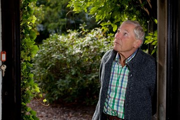 schlanker Senior mit weißem Haar und blauen Augen steht in einer Gartentür, guckt nach oben und...