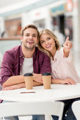 attractive young woman pointing by finger to smiling boyfriend at table with disposable cups of coffee in cafe
