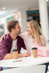 smiling couple with shopping bag looking at each other at table with disposable cups in cafe