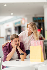 shocked young man looking at shopping bags at table with paper cup of coffee while his girlfriend standing near in cafe