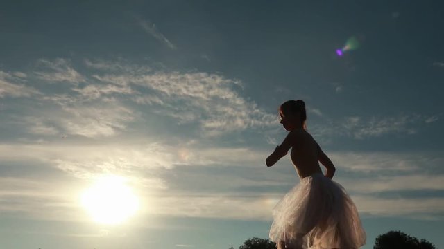 Ballerina in white transparent dress dances on background of sunset and blue sky in the evening. Backlight. Bottom view. 