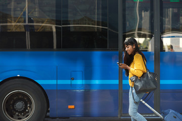 smiling young Indian woman walking with suitcase and cellphone by bus station © mimagephotos