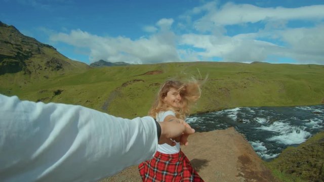 POV of man holding hand of girlfriend, woman or partner, leading towards amazing epic view of waterfall and mountain river. Beautiful blond girl looks back smiling, adventures and relationship goals