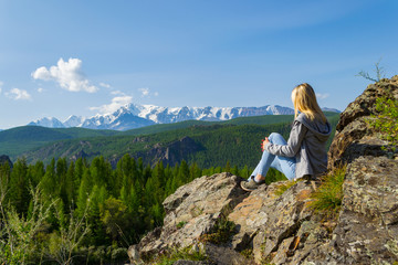 Young blonde girl on a rock on a sunny warm autumn summer day resting while traveling near a green tree on the background of landscape snow-capped mountain top and a blue sky with clouds in Altai