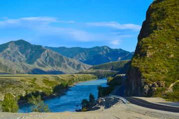 A new winding asphalt black road with a yellow dividing strip between the rocks and the Katun River in the Altai mountains under a clear blue sky with clouds and a field covered with green grass