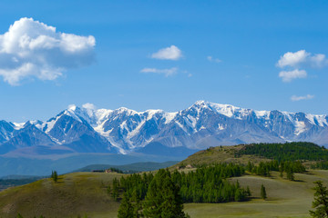 Landscape of snow-capped mountains, with green hilly valley a fresh summer day under a blue sky with white clouds and sun rays in Altai mountains