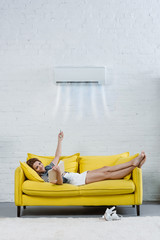 Fototapeta na wymiar relaxed young woman reading book on couch and pointing at air conditioner hanging on wall with remote control
