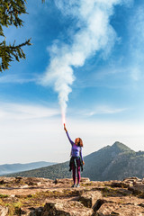 A woman in the wild mountains gives a distress signal SOS using Falsch feuer torch from which comes a bright flame and orange smoke, Concept of emergency situation during hike in the woods
