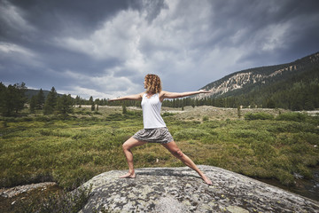 a young woman with curly hair doing yoga on top of a giant boulder in the mountains