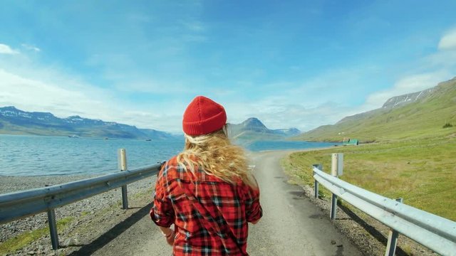 Camera in slow motion follows young blond woman walk on empty gravel road in amazing epic landscape in iceland, inspiration for travel destination or blogger content, looks in camera smiling