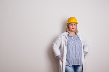 Portrait of an attractive overweight woman with yellow helmet and lab coat in a studio.