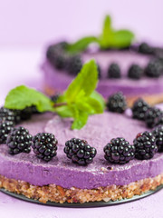 Raw blackberry dessert decorated with fresh ripe forest berries and green mint leaves on pastel violet background.